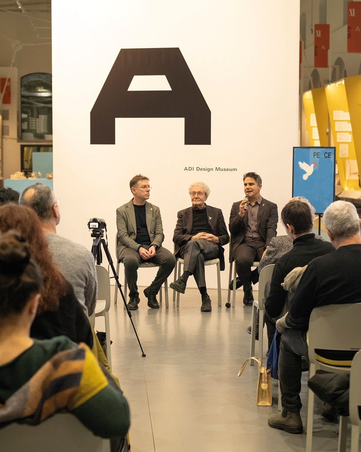 Tre relatori seduti davanti al pubblico durante un incontro all’ADI Design Museum, uno parla al microfono.